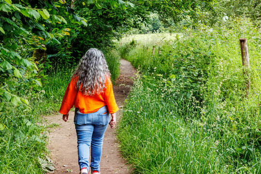 Adult hiker woman walking calmly on a trail, back to camera, meadow covered with wild green grass, casual clothes, jeans and orange blouse, sunny summer day in Namur province, Wallonia, Belgium