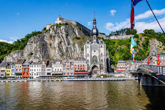 Cityscape with citadel on top of rocky mountain and collegiate church of Our Lady of Dinant, bridge with waving flags, buildings facing river Meuse, sunny summer day in Namur, Wallonia, Belgium