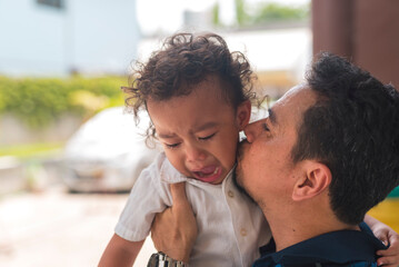 A dad tries to calm down his scared or hurt young son by picking him up and kissing him, telling it is alright.