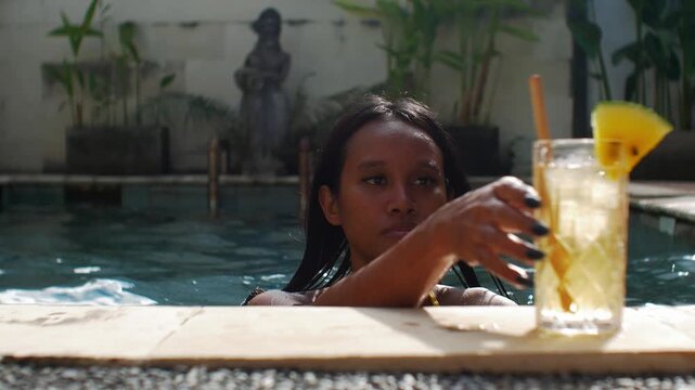 Asian model in bikini swims to poolside with refreshing tropical cocktail. She enjoys the cool water on a hot and sunny summer pool day on Bali Island.(30 to 24fps slow motion, focus pull, bokeh)