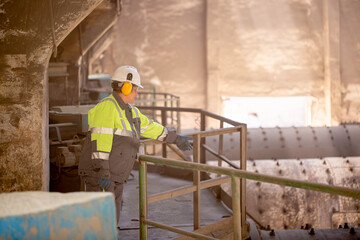 A worker in a helmet and equipment stands against the backdrop of a cement plant workshop. Work technology, cement production. People working in heavy industry. © Vera