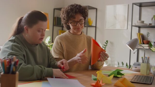 Girl with Down syndrome sitting at desk and crafting origami with help of female teacher during art class