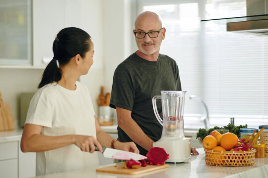 Portrait of smiling couple preparing smoothie in modern kitchen while chatting. Man blending ingredients, woman cutting dragonfruit. Modern kitchen with various fruits - Powered by Adobe