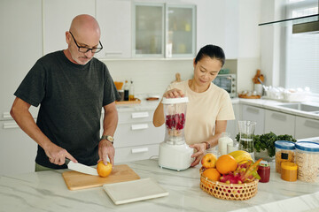 Senior man slicing orange while woman blending fruits for a smoothie on kitchen countertop. Kitchen has modern appliances and fresh ingredients visible