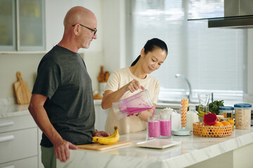Asian woman blending fruit while Caucasian man looks on in modern kitchen. Preparing a fruit smoothie atop island counter