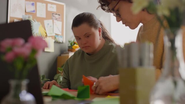 Girl with Down syndrome sitting at desk, listening attentively to female teacher and folding paper while learning origami during arts and crafts lesson