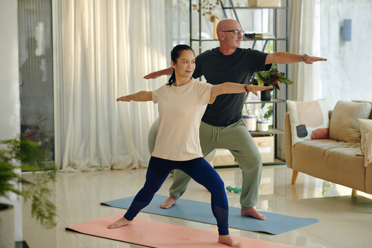 Woman and man practicing yoga poses on mats in living room surrounded by furniture and houseplants, focusing on balance and posture without using additional props and equipment