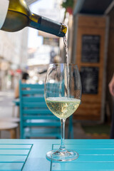 A close-up of a hand holding a glass of white wine in an outdoor cafe, with a blurred background featuring people and outdoor seating in a lively atmosphere.