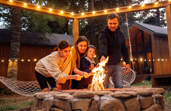 A young married couple with children roasts marshmallows on the fire. A man with his wife, son and daughter are having fun in the backyard of the house.