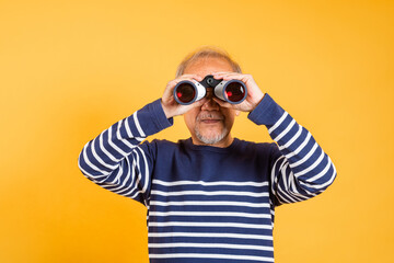 Portrait Asian smiling old man looking camera through binoculars studio shot isolated yellow background, senior man pensioner feeling surprised using binocular