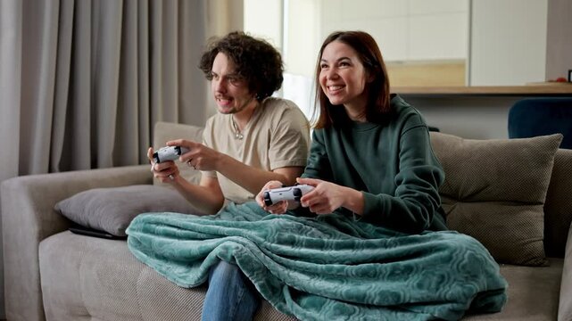 Confident brunette guy with curly hair plays video games using a joystick with his girlfriend in a green jacket but the girl wins him on the sofa in a modern apartment