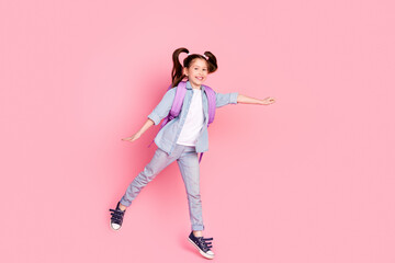 Full size photo of positive lovely small girl dressed denim shirt rucksack on shoulders jumping to empty space on pink color background