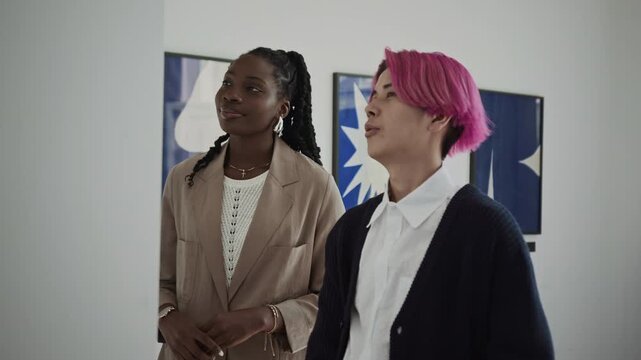 Young Asian guy with pink hair smiling and discussing artwork with African American girlfriend while visiting gallery exhibition together