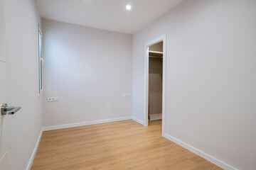A simple, empty room featuring wooden flooring and an open closet, accented by a window and clean white walls, providing a bright and welcoming atmosphere.