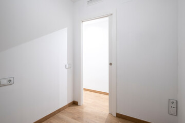 A minimalistic unfurnished white room with clean walls and a smooth wooden floor, featuring an open sliding white door leading to another, well-lit space.