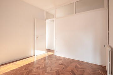 An empty room featuring herringbone patterned wood flooring with a modern minimalistic design, white walls, and a door partially open letting in natural light.