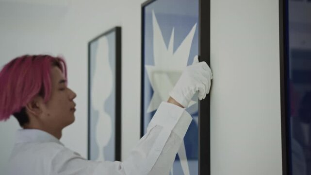 Young Asian gallery worker in white gloves hanging framed contemporary artwork on wall while preparing for exhibition opening