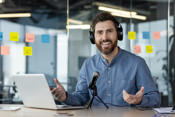 Portrait of a young man sitting in the office at the desk wearing headphones in front of a microphone and a laptop, talking and looking at the camera with a smile, gesturing with his hands