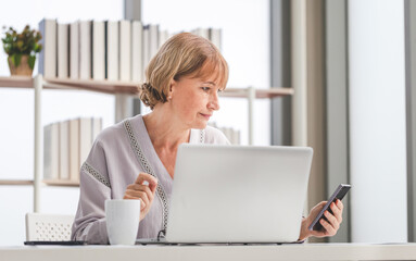 A middle-aged woman works at the table and looking at the smartphone, Woman using smartphone and laptop