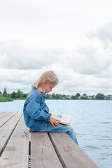 Girl reads a book while sitting on the pier. Education and learning concept. Vertical orientation.