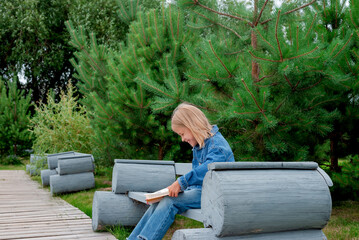 Fototapeta premium Laughing girl reading a book sitting on a park bench. Education and learning concept.