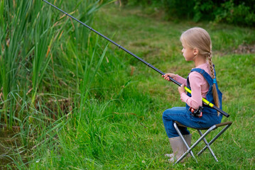 Little girl catches fish with a fishing rod on the edge of a pond while sitting on a chair.