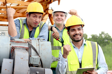 Cheerful worker team with safety vest and helmet raising hand up at construction building site. Young labor hold tablet with senior engineer and elderly foreman raise fists to celebrate after job done