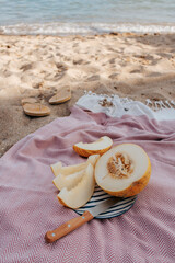 A pink blanket lies on the sand by the sea. On it is a cut melon on a striped plate and a knife...