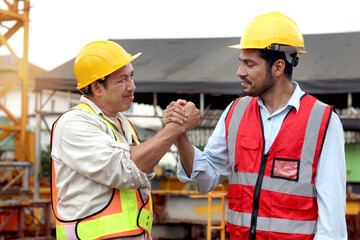 Happy engineers with safety vest and helmet have strong handshake at building site. Senior elderly Asian shakes hands with young worker during works together at construction site. teamwork and unity.