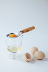 in a glass cup raw egg white, on top is an egg separator with yolk. next to it is a shell and eggs
