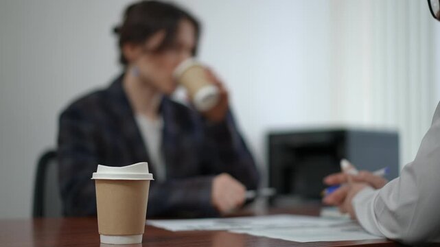 In the foreground, a disposable paper cup with coffee stands on a desk in an office. In the background in a weak defocus, a female manager in a blazer drinks coffee from a paper cup and talks to an