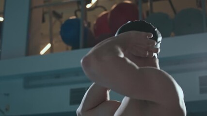 Tilt up slowmo shot of Caucasian male swimmer with black tattoo on his rib putting on swimming cap before start of race in indoor pool