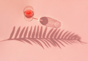 Creative summer scene wine glass with red wine and shadow of palm leaf. Flat lay on pink background.