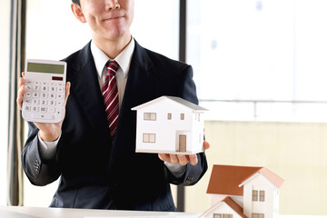 住宅模型と電卓を持ち悩むスーツ姿のアジア人男性のビジネスパーソン