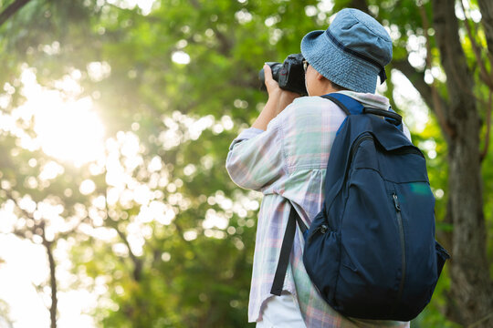 Portrait of Asian mature woman holding a camera and backpack behind her back, an Asia active senior woman enjoying nature in park. Standing on a trail in a forest outdoors. Enjoying active travel trip