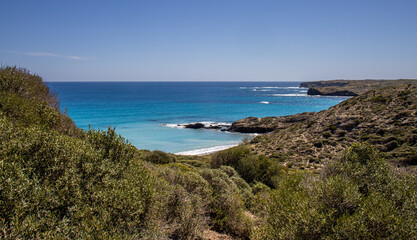 Panoramic view of the idyllic cove of Presili on a sunny day. The Mediterranean Sea displays crystal-clear turquoise and blue hues, contrasting with the golden sand of the beach. Menorca, Spain