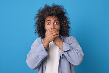 Young Indian man with curly hair stands on blue backdrop, his eyes wide open in astonishment. He covers his mouth with both hands, conveying mix of shock and disbelief