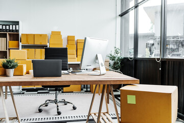 A wooden desk in an SME office, with a laptop, desktop computer, and a parcel cardboard box. The workspace is organized for efficient management, logistics, and order processing.