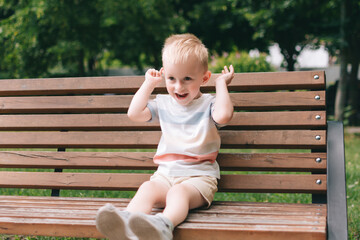 A boy with fair hair makes faces while sitting on a bench in a summer park.