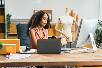 An African American woman with an afro works at a wooden desk, equipped with a laptop, desktop computer, and parcel cardboard boxes, focusing on SME logistics, inventory, and online orders.