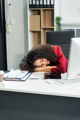 A hardworking African American woman with an afro sits tired at a wooden desk, suffering from office syndrome, as she works on her business finance startup in a busy office workplace.