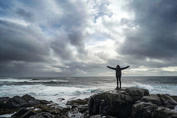 a person standing on a rocky shoreline with arms outstretched, enjoying the serene view of the sea under a cloudy sky