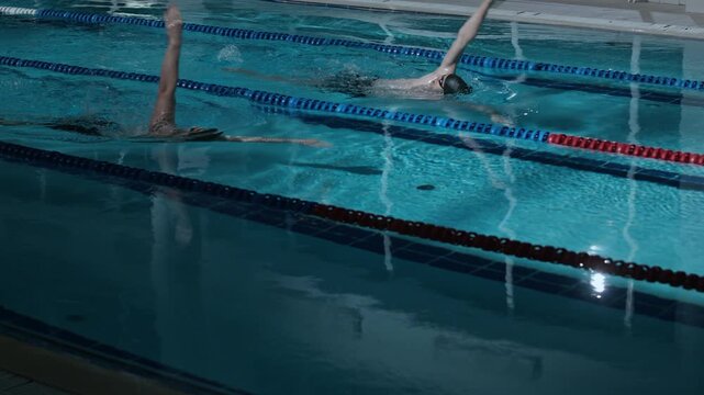 Two athletic Caucasian men practicing backstroke swimming in lane pool while training together indoors