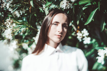 A true tilt-shift portrait of a woman in a white shirt is standing in front of lush green foliage and white flowers. The photo emphasizes her serene expression and the detailed background