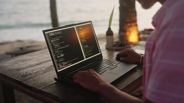 Web developer works on laptop in beachside cafe at sunset, programming with code on screen. Ocean waves, warm light, serene backdrop as male programmer codes software near sea. Slow motion.