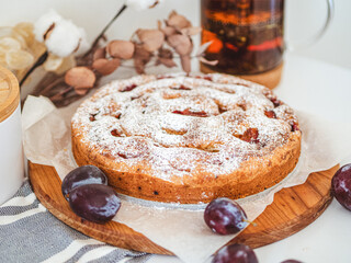 Homemade plum pie with powdered sugar and cinnamon on a wooden board near a teapot and dried flowers.