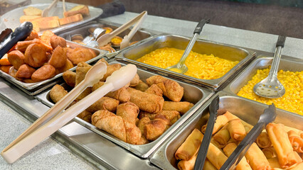a buffet-style food setup with trays of various fried items, such as spring rolls and breaded snacks, alongside macaroni and cheese, and corn