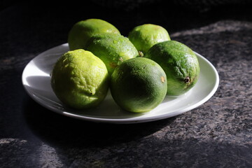Una toma de cerca de varios limones verdes con agua  en un plato de color blanco en la cocina