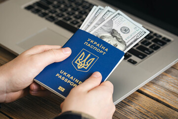 Ukrainian biometric passport and dollar bills in the hands of a man against the background of a laptop.