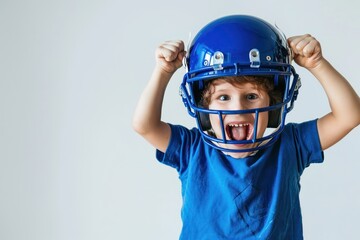 Young child celebrates wearing oversized American football helmet and blue jersey on white background. Arms raised high in excitement, face lit up with wide smile.